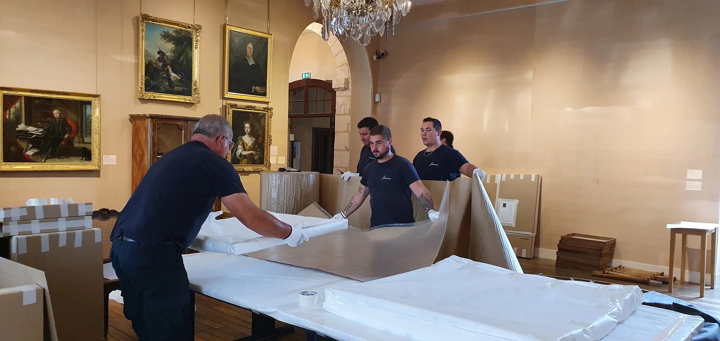 Des déménageurs Jumeau en gants blancs étendent une grande feuille de protection sur une table d’emballage, devant des tableaux à cadre doré