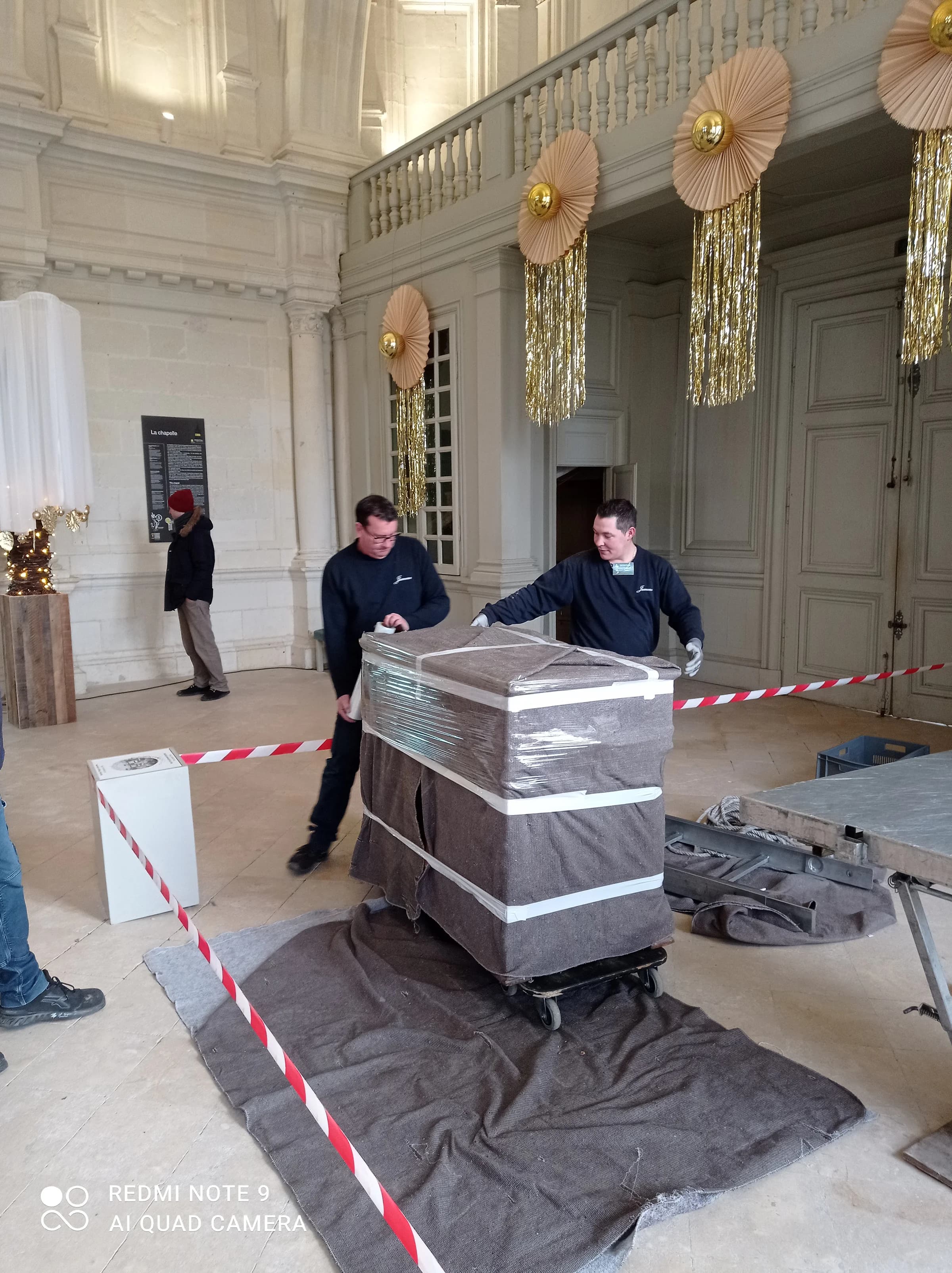 Deux déménageurs Jumeau poussent sur un chariot une caisse de transport d’œuvre d’art, sous housse et film, dans une grande salle