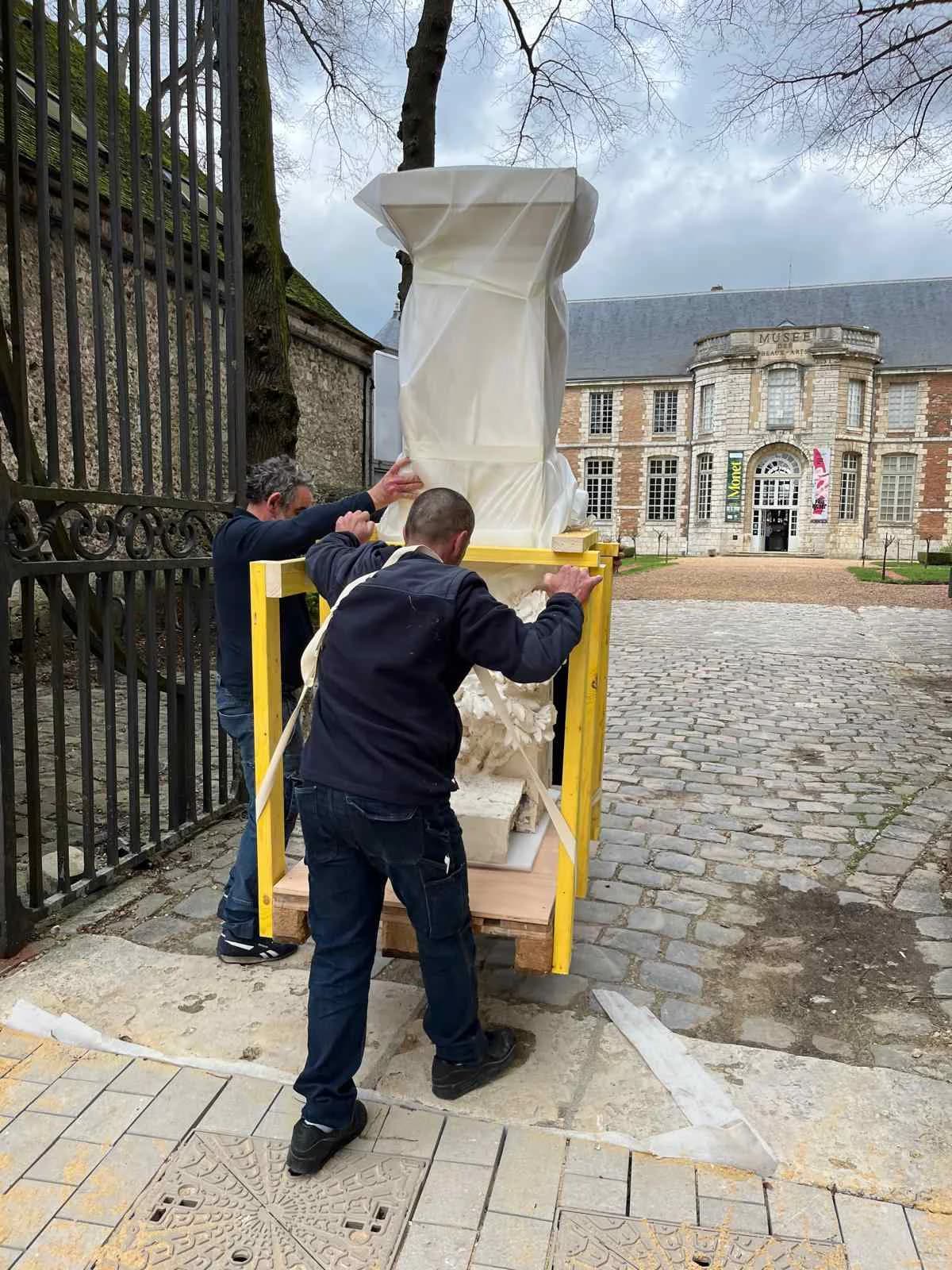Manutention d’une sculpture en partie emballée sous film, sanglée dans un cadre de bois sur palette et poussée par deux hommes, devant le Musée des Beaux-Arts