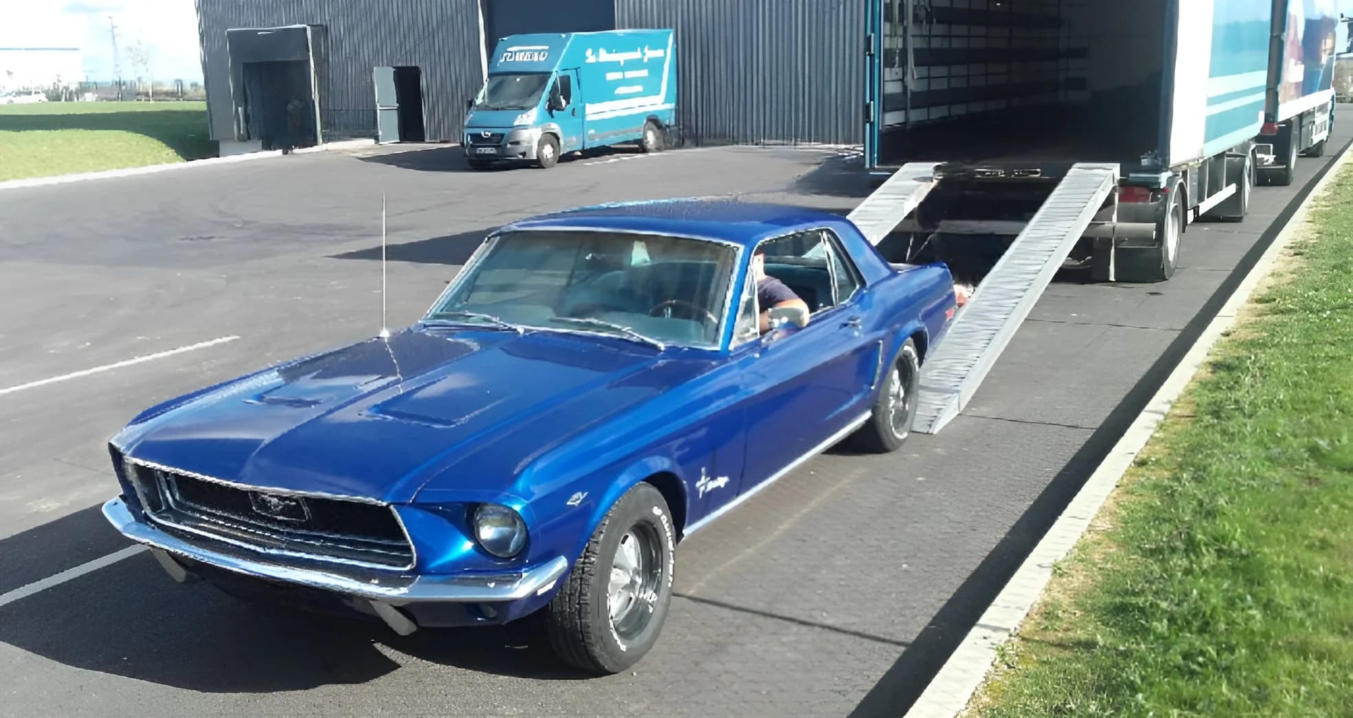 Une Ford Mustang ancienne, bleue, au pied des rampes d’une remorque, sur le parking d’un entrepôt