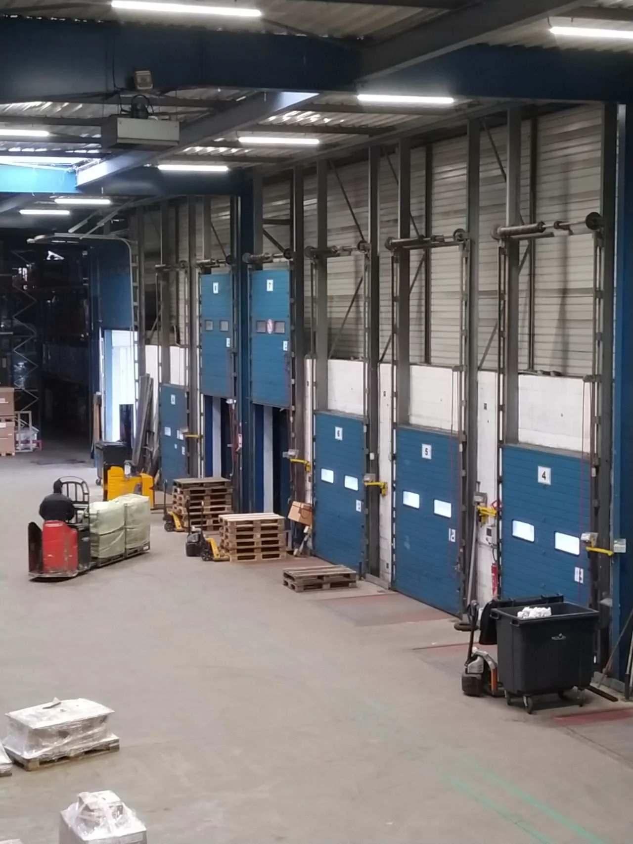 Vue plongeante sur une halle de quais de chargement et sa rangée de portes sectionnelles bleues, dont certaines sont levées. Au sol, un cariste sur un transpalette électrique rouge chargé de colis sous film, des palettes vides, des transpalettes et des bacs roulants.
