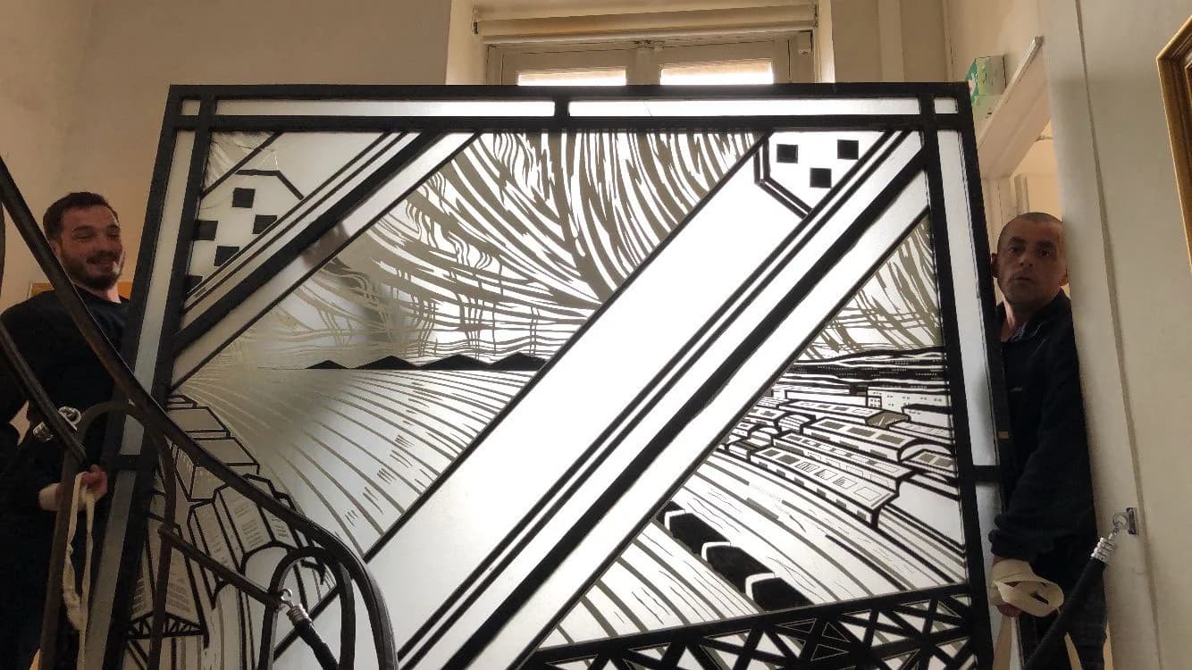 Deux déménageurs portent un grand vitrail au dessin noir et blanc, dans un escalier à rampe de fer forgé