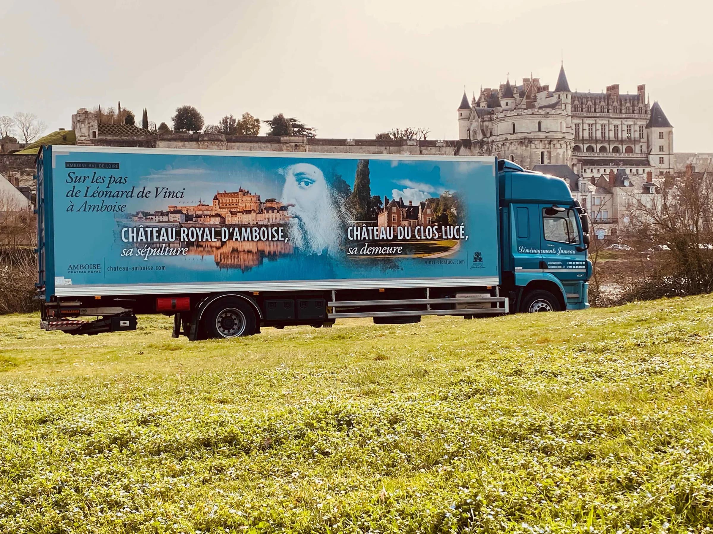 Un camion des Déménagements Jumeau, la caisse habillée d’un visuel du château royal d’Amboise et du château du Clos Lucé, dans un pré au pied d’un château