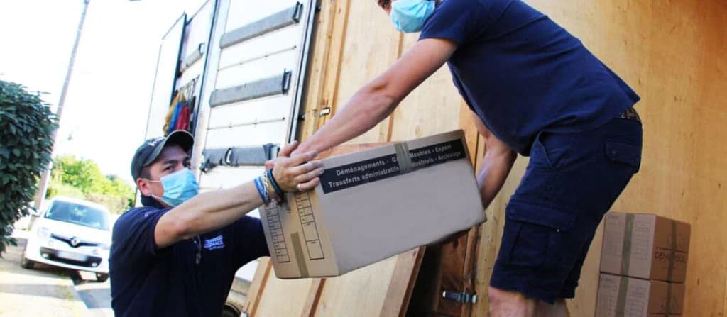 Deux déménageurs masqués se passent un carton à l’arrière d’un camion, une voiture garée derrière eux