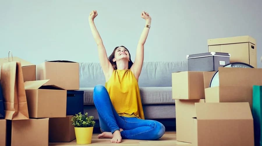 Une jeune femme assise par terre au milieu de cartons lève les bras de joie, devant un canapé
