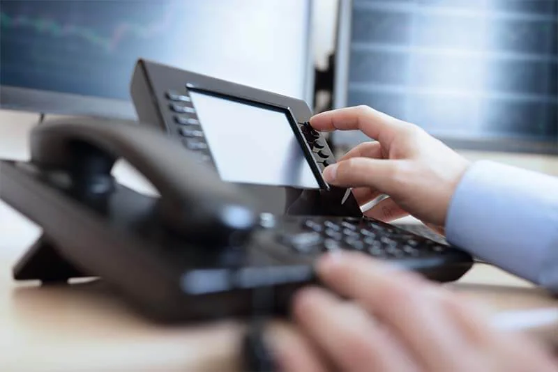 Une main compose un numéro sur un téléphone de bureau, devant deux écrans