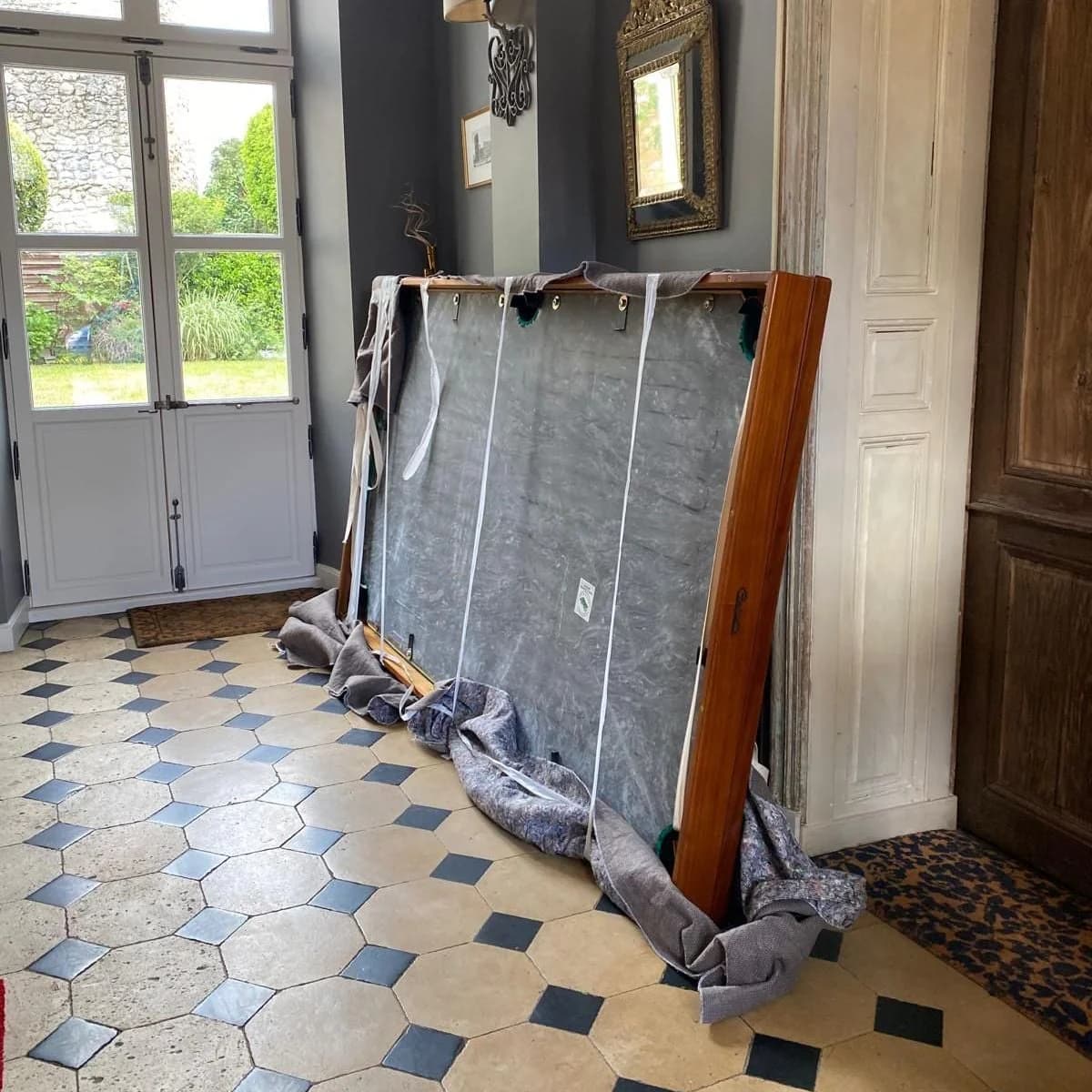 Un billard emballé sous des couvertures, posé debout dans un hall au carrelage ancien
