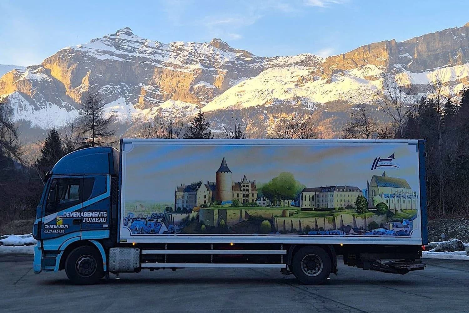 Un camion des Déménagements Jumeau habillé d’une peinture de château, au pied de montagnes enneigées, en Italie