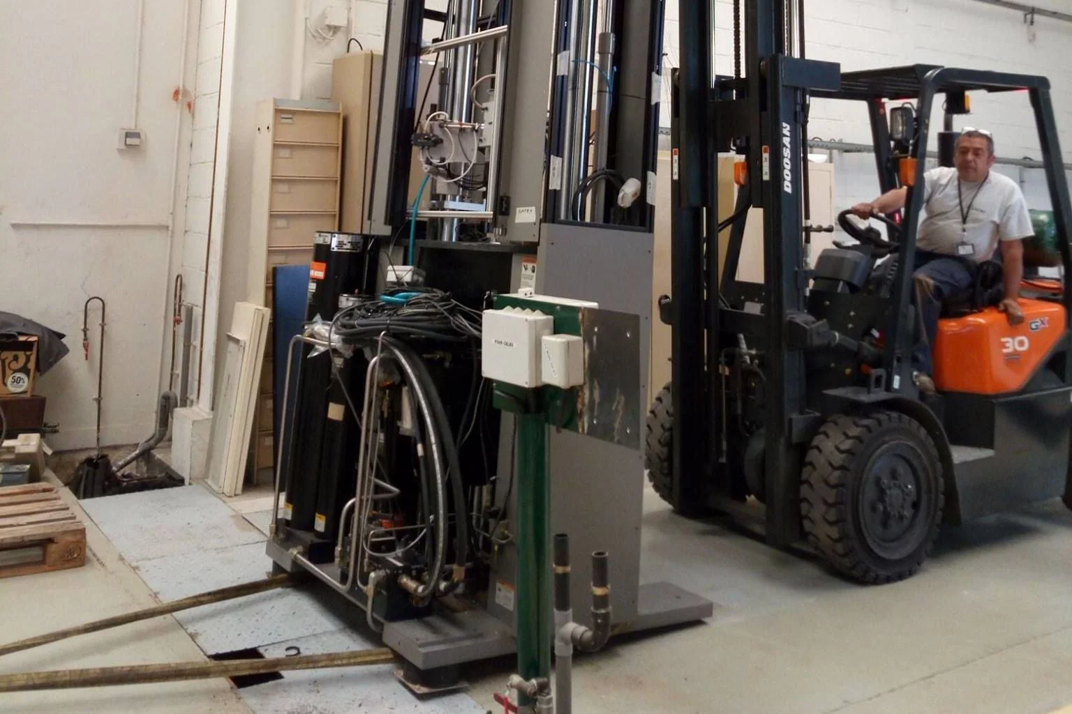 Un cariste au volant d’un chariot élévateur, à côté d’une machine industrielle sur son socle, dans un atelier
