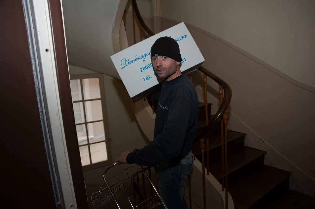 Un déménageur monte un escalier, un carton des Déménagements Jumeau sur l’épaule