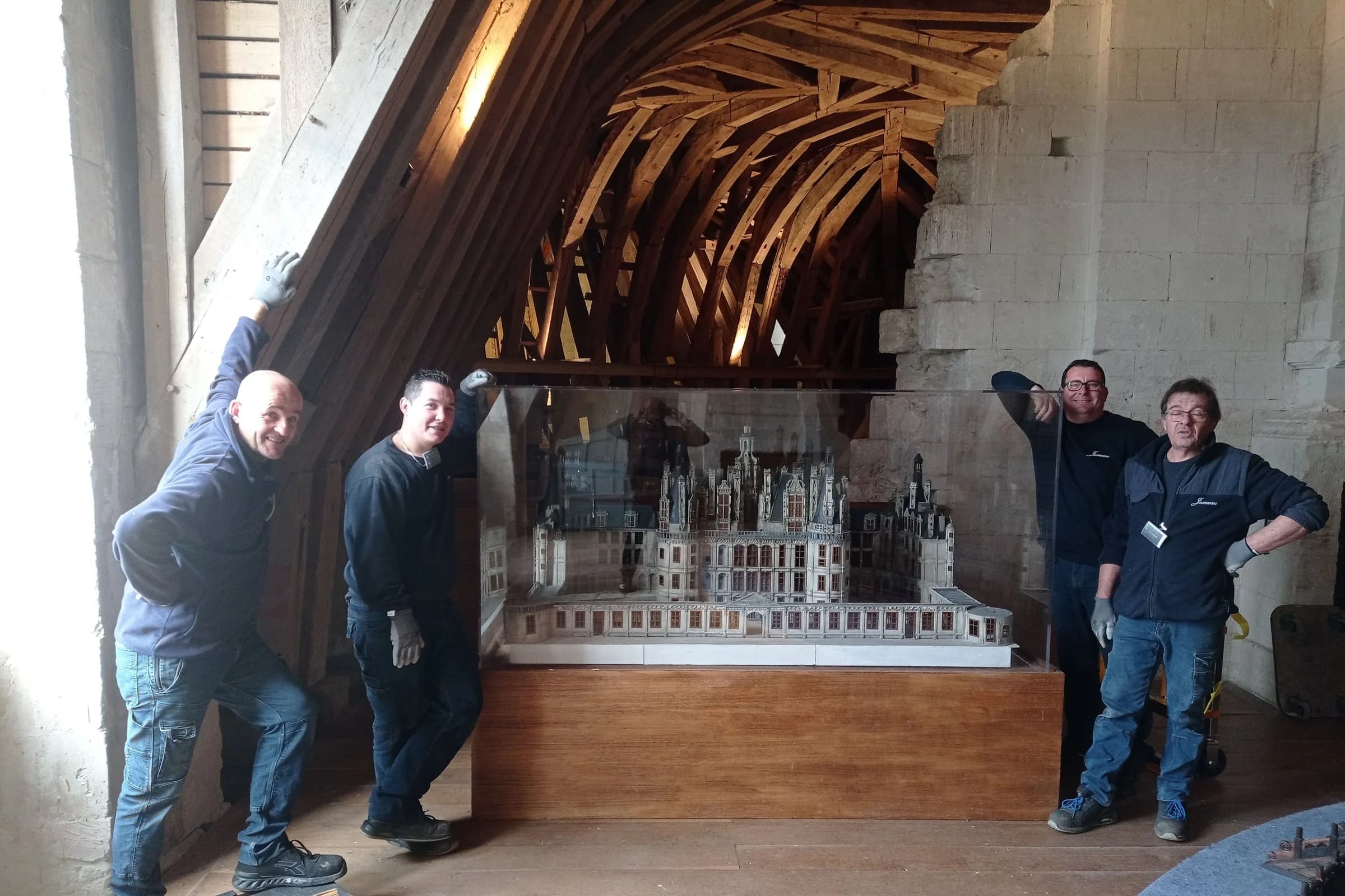 Quatre déménageurs de Jumeau posent autour d’une maquette du château de Chambord, sous une charpente