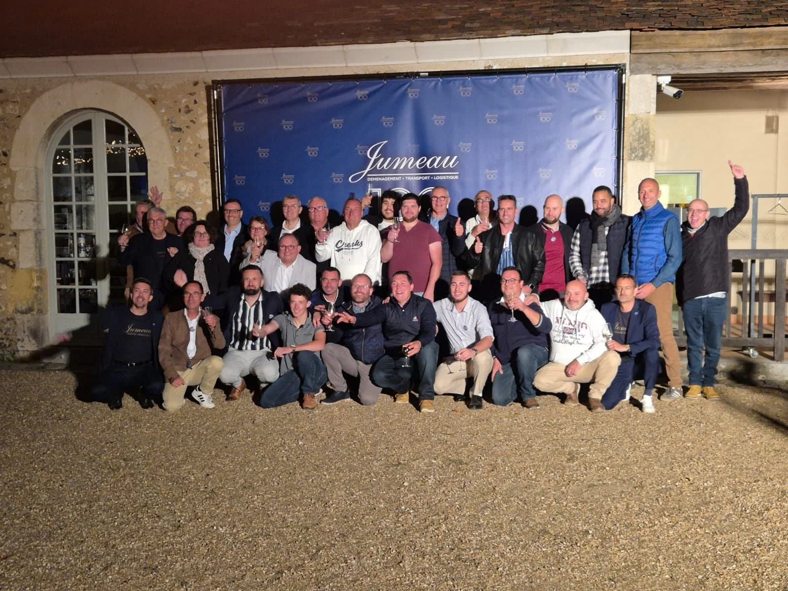 Une photo de groupe d’une quarantaine de personnes devant une bâche au nom de Jumeau