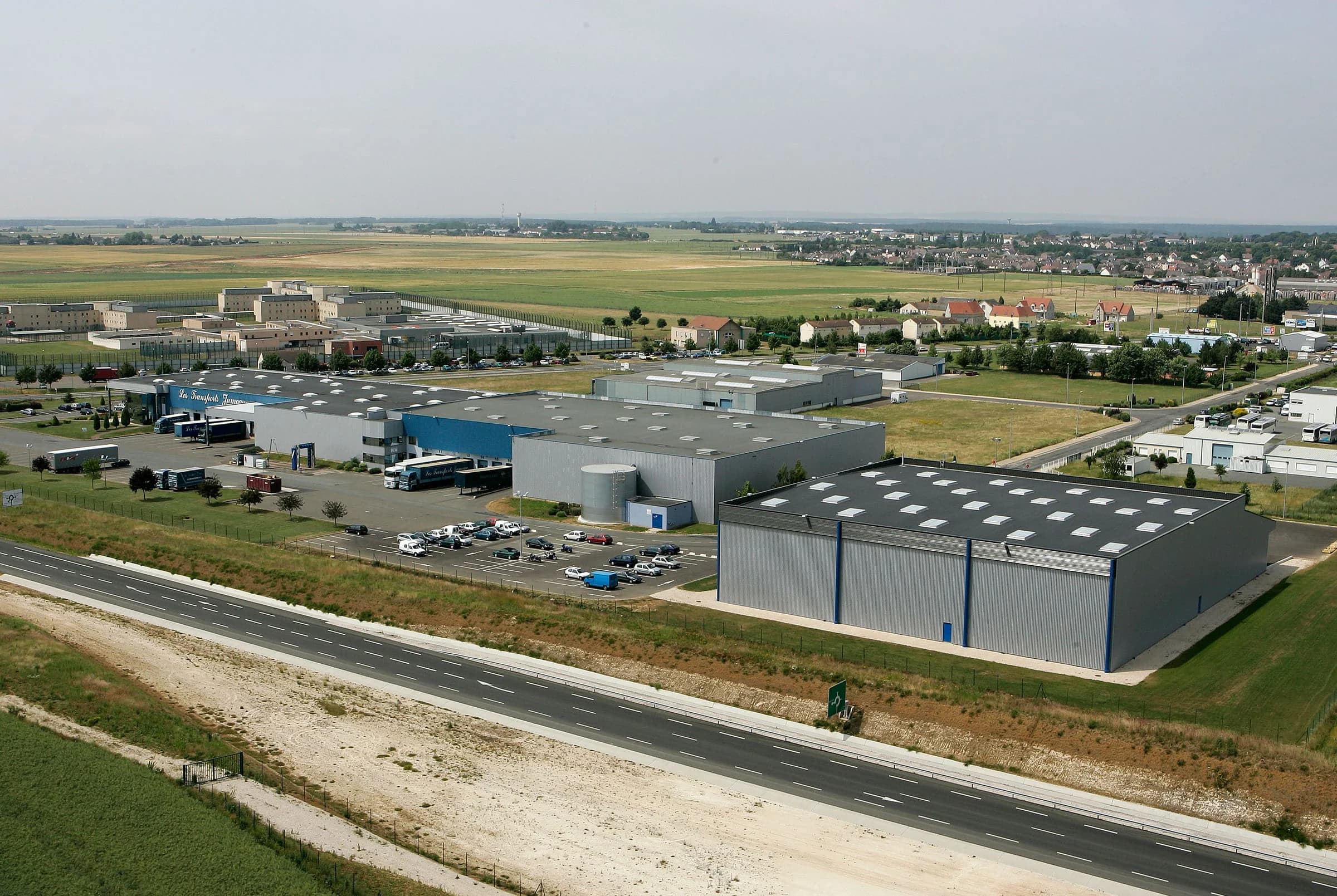 Vue aérienne de grands entrepôts gris le long d’une route, au milieu des champs