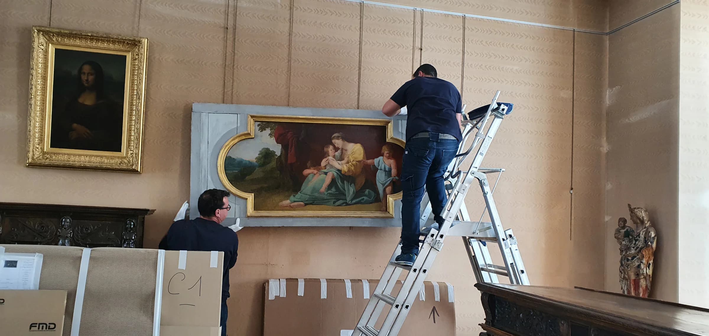 Deux hommes en gants blancs manipulent un grand tableau encadré dans une salle ancienne, l’un sur un escabeau