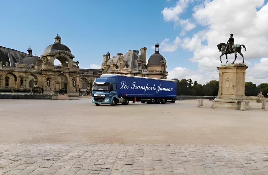 Un semi-remorque bleu à l’enseigne des Transports Jumeau dans la cour pavée d’un château, près d’une statue équestre