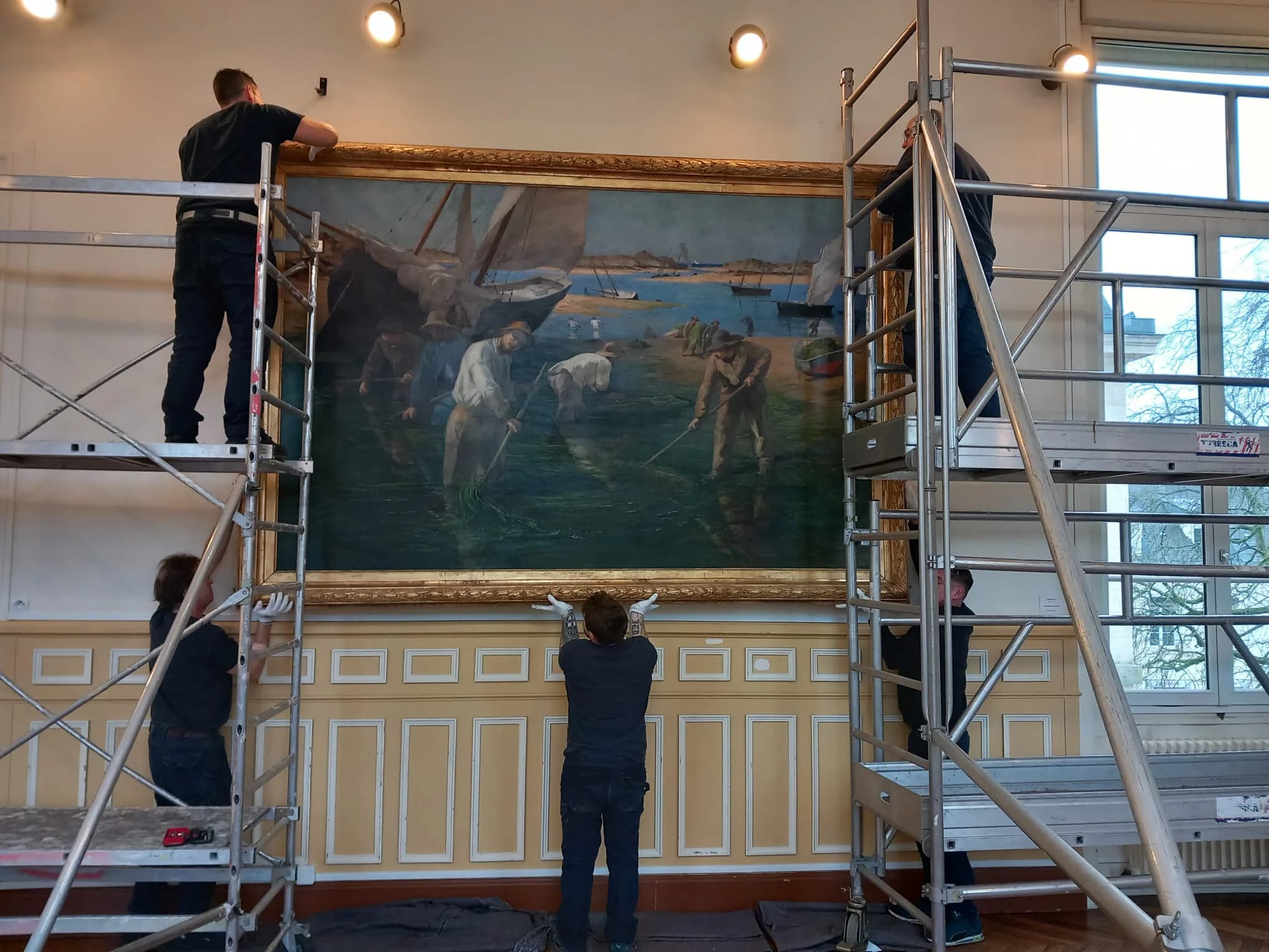 Une équipe en gants blancs accroche un grand tableau à cadre doré, depuis deux échafaudages