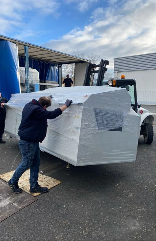 Un déménageur accompagne une machine emballée sous film, portée par un chariot élévateur, au pied d’un camion bâché