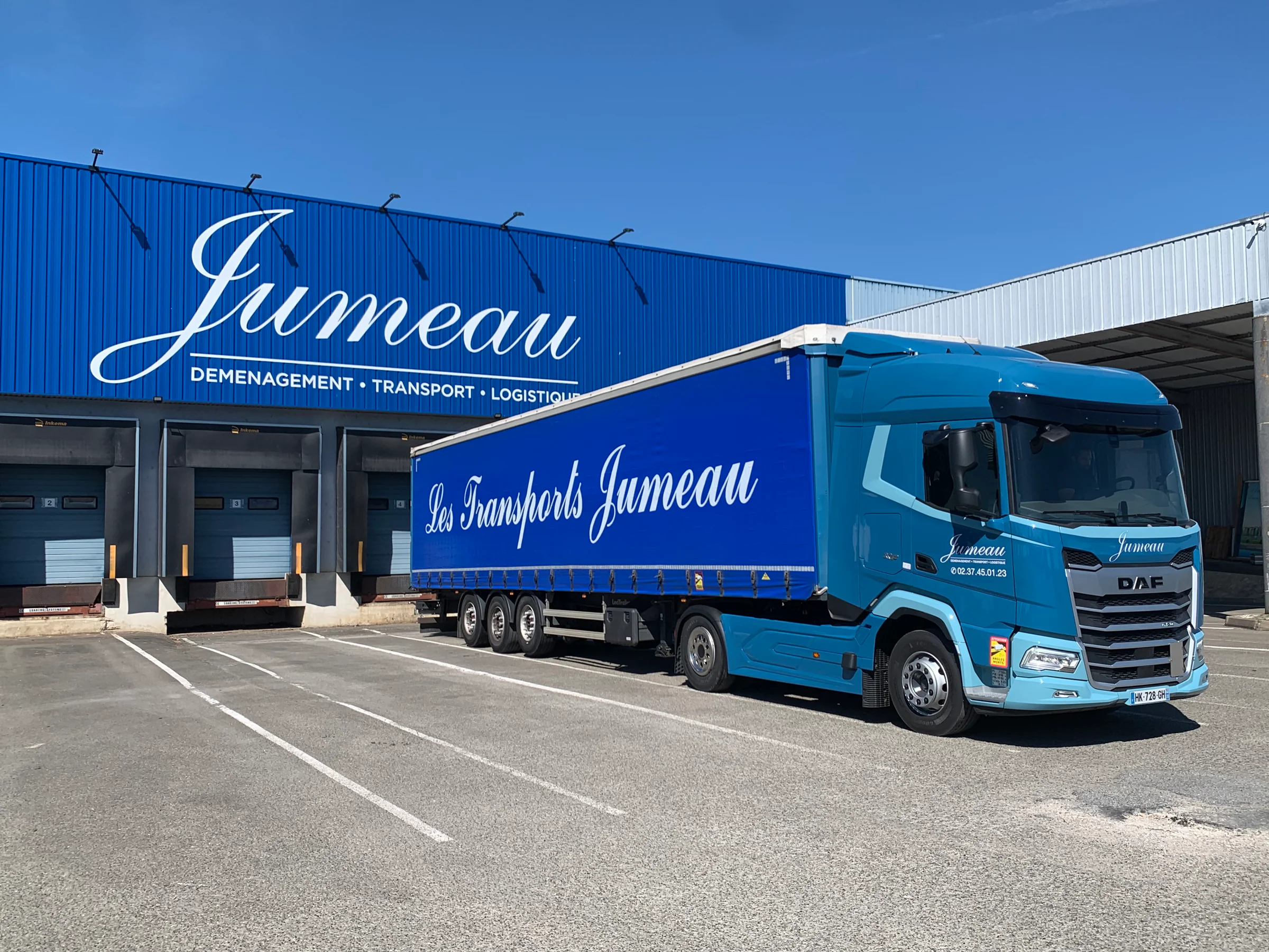 Une semi-remorque bâchée « Les Transports Jumeau » devant les quais de l’entrepôt Jumeau, « Déménagement • Transport • Logistique »