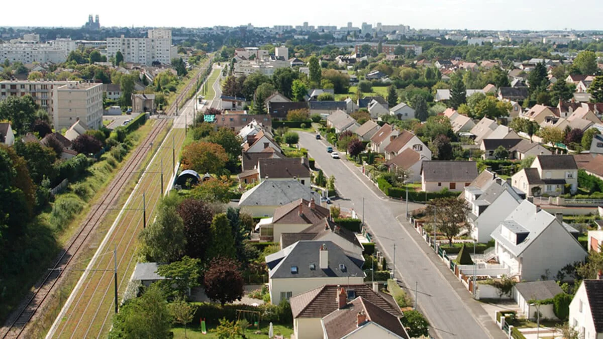 Vue aérienne d’un quartier pavillonnaire, entre une rue et une voie ferrée, des immeubles à l’horizon