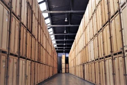 Stockage de caisses en bois empilées en hauts murs le long d’une allée, sous la charpente métallique éclairée d’un hangar