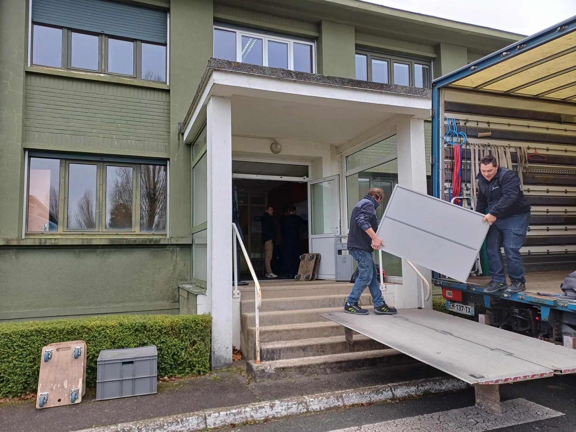 Deux déménageurs chargent un grand panneau sur le hayon d’un camion, devant l’entrée d’un bâtiment