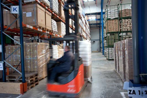 Dans une allée d’entrepôt, un cariste conduit un chariot élévateur rouge, flou de mouvement. À gauche, des racks chargés de cartons sur palettes. À droite, des sacs dans des casiers grillagés verts et des palettes de cartons posées au sol.