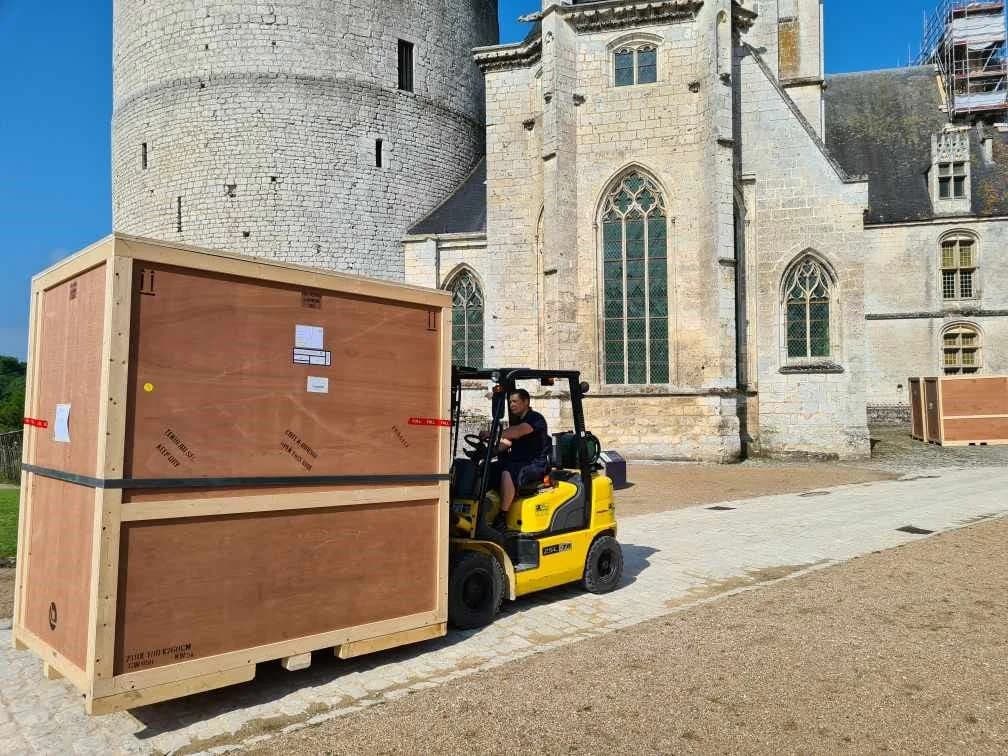 Un chariot élévateur porte une grande caisse de transport en bois devant un château, d’autres caisses attendent au fond