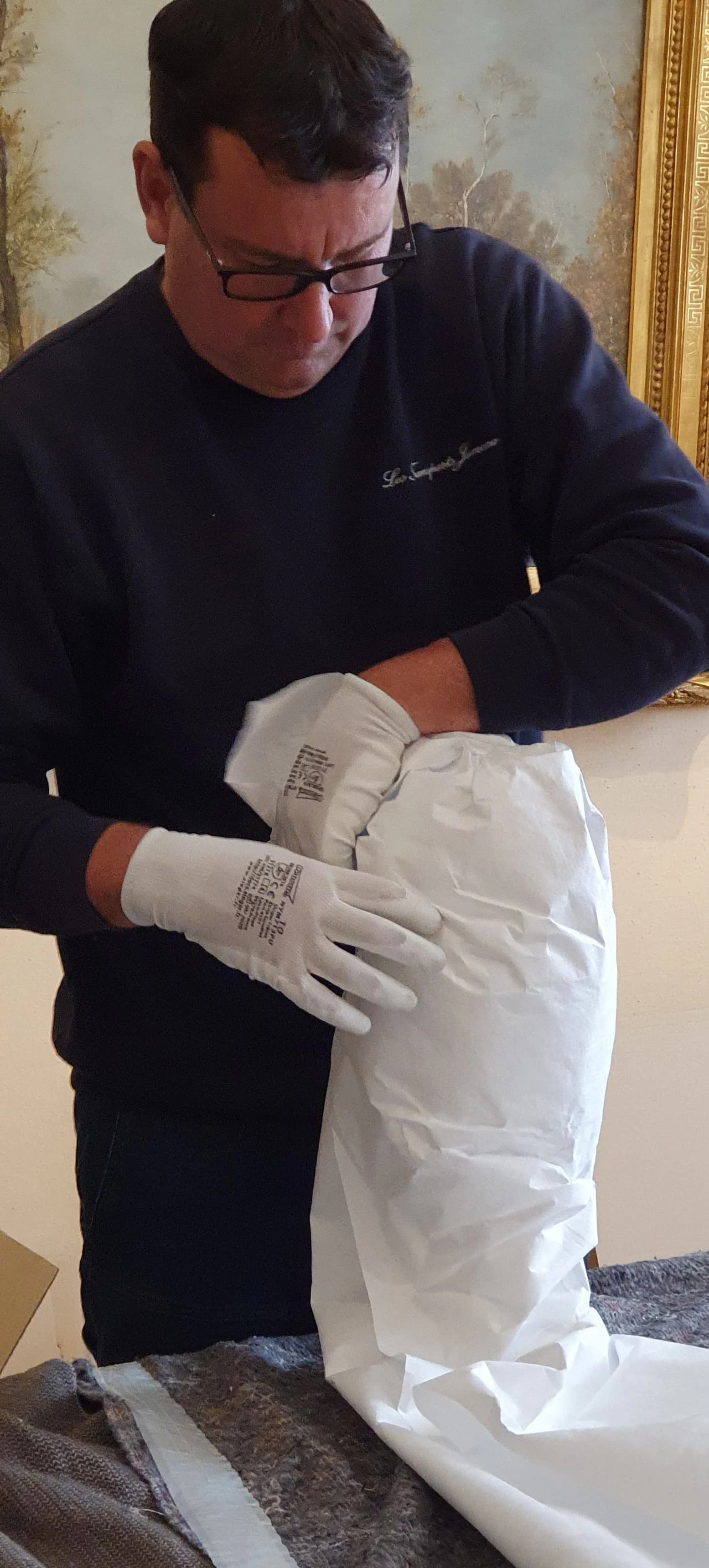 Un déménageur Jumeau en gants blancs glisse un objet dans une housse de protection, devant un tableau à cadre doré