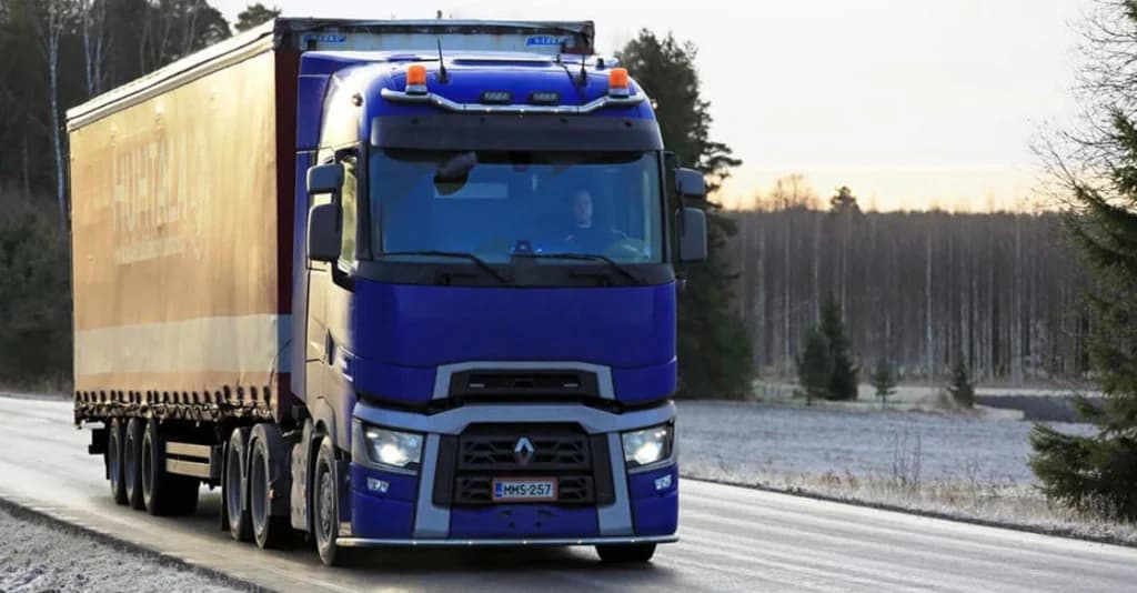 Un semi-remorque à cabine bleue roule sur une route givrée bordée de bouleaux, sous un ciel d’hiver