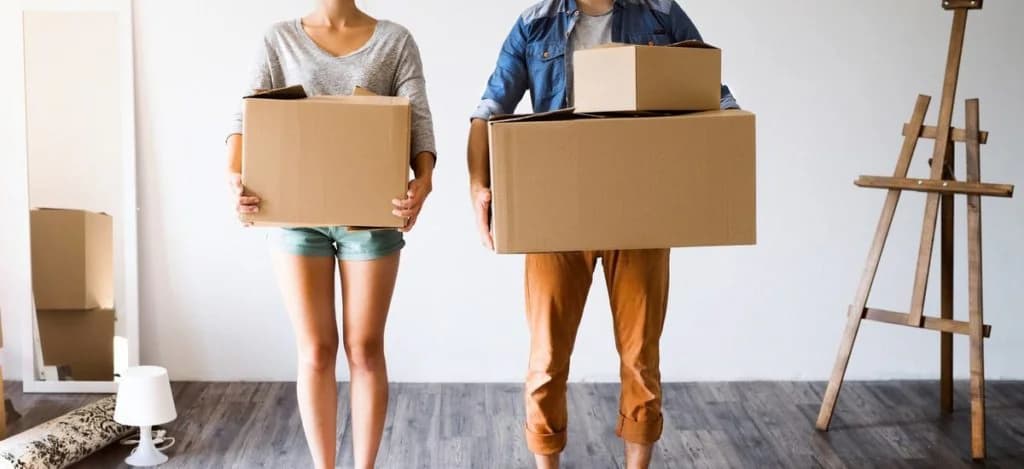 Un couple, visages hors cadre, tient des cartons dans une pièce vide, près d’un miroir et d’un chevalet