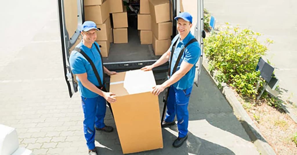 Deux déménageurs souriants, en bleu et casquette, tiennent un grand carton devant un fourgon rempli de cartons