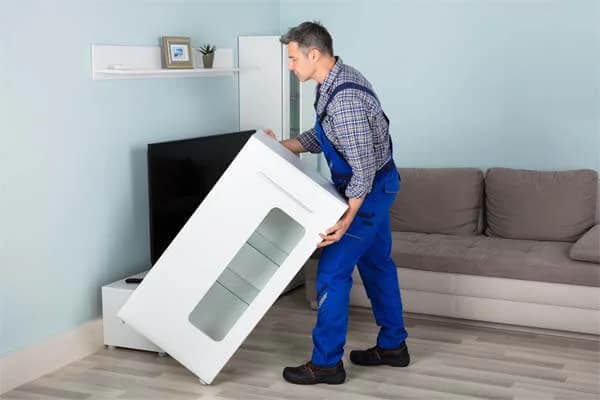 Un homme en salopette bleue soulève un meuble blanc dans un salon, près d’un canapé et d’un téléviseur
