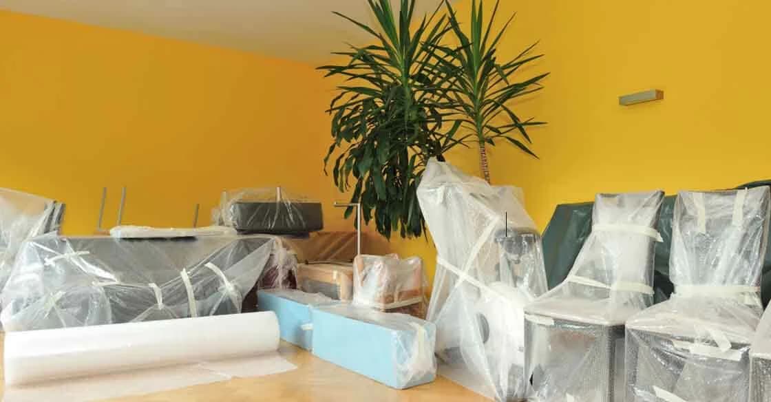 Chaises et meubles emballés sous film plastique et papier bulle, dans une pièce aux murs jaunes