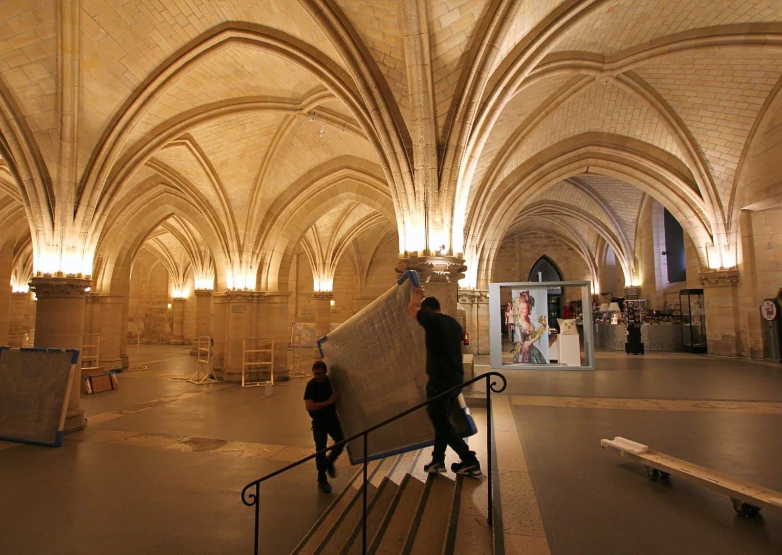 Deux déménageurs montent une grande caisse plate dans la salle gothique voûtée de la Conciergerie