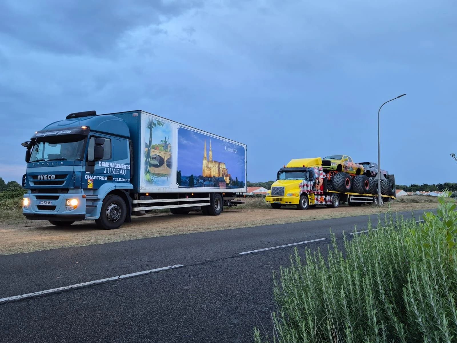 Un camion des Déménagements Jumeau sur une route, suivi d’un camion américain jaune qui porte un monster truck