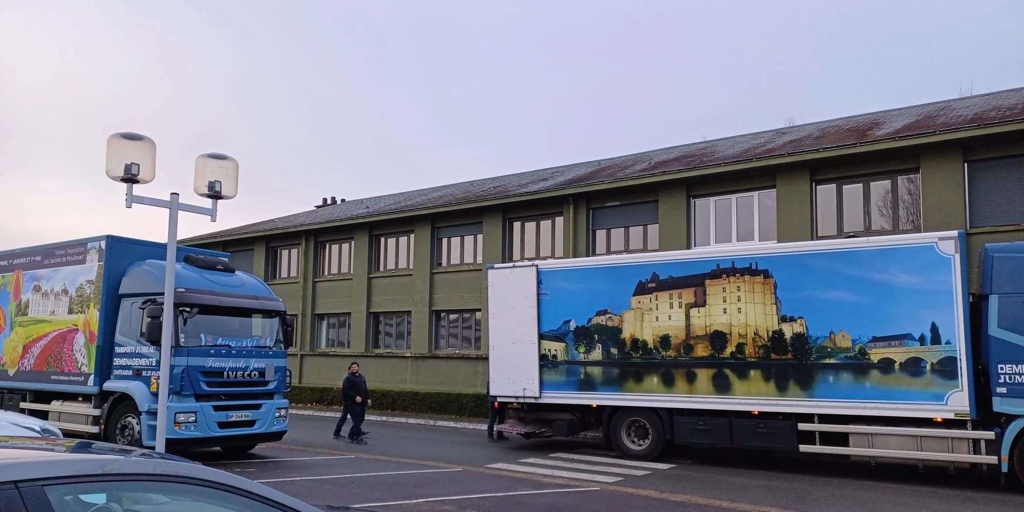 Deux camions de Jumeau à Châteaudun, l’un habillé d’une peinture de château au bord de l’eau, garés devant un long bâtiment