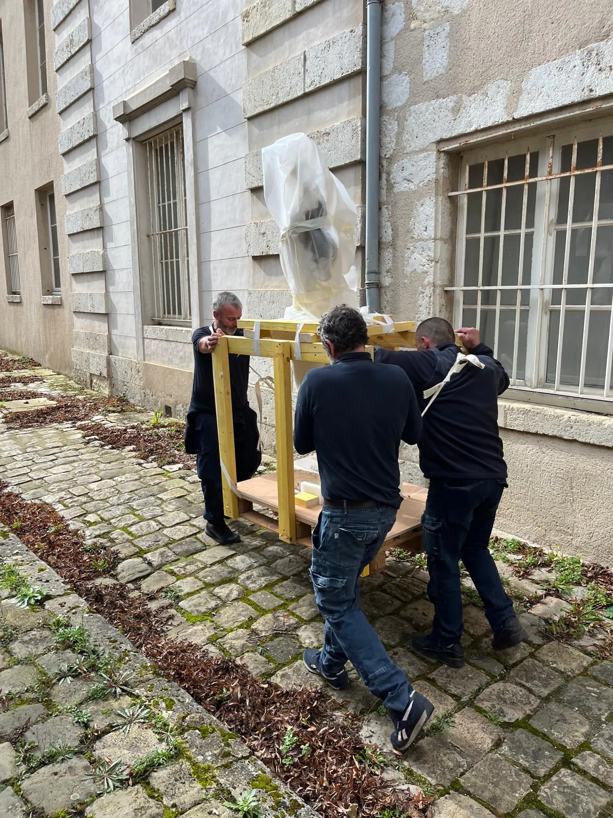 Trois déménageurs portent une statue emballée dans un cadre de bois, dans une cour pavée