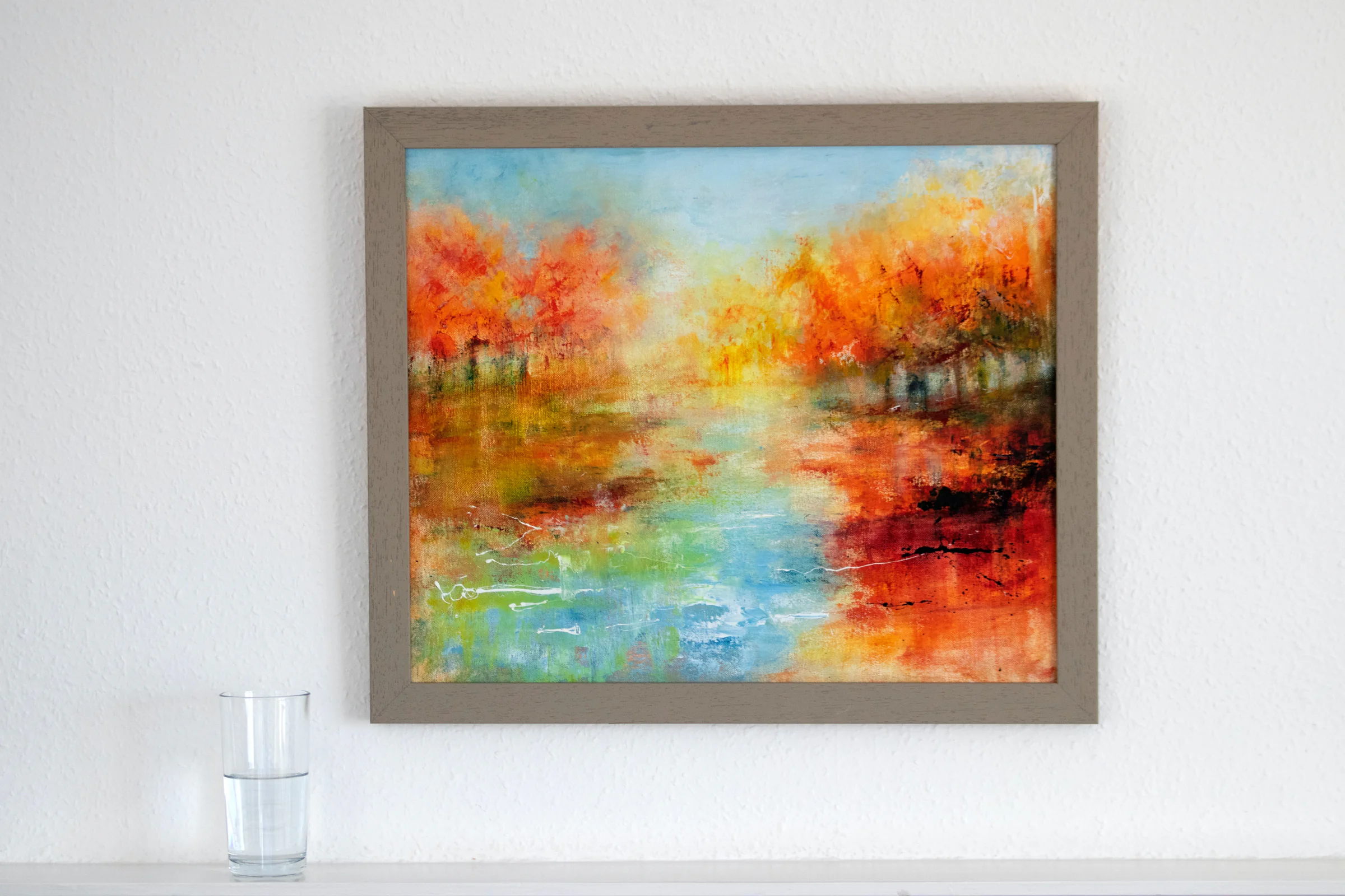 Un tableau encadré, une rivière bordée d’arbres aux couleurs d’automne, accroché au mur près d’un verre d’eau